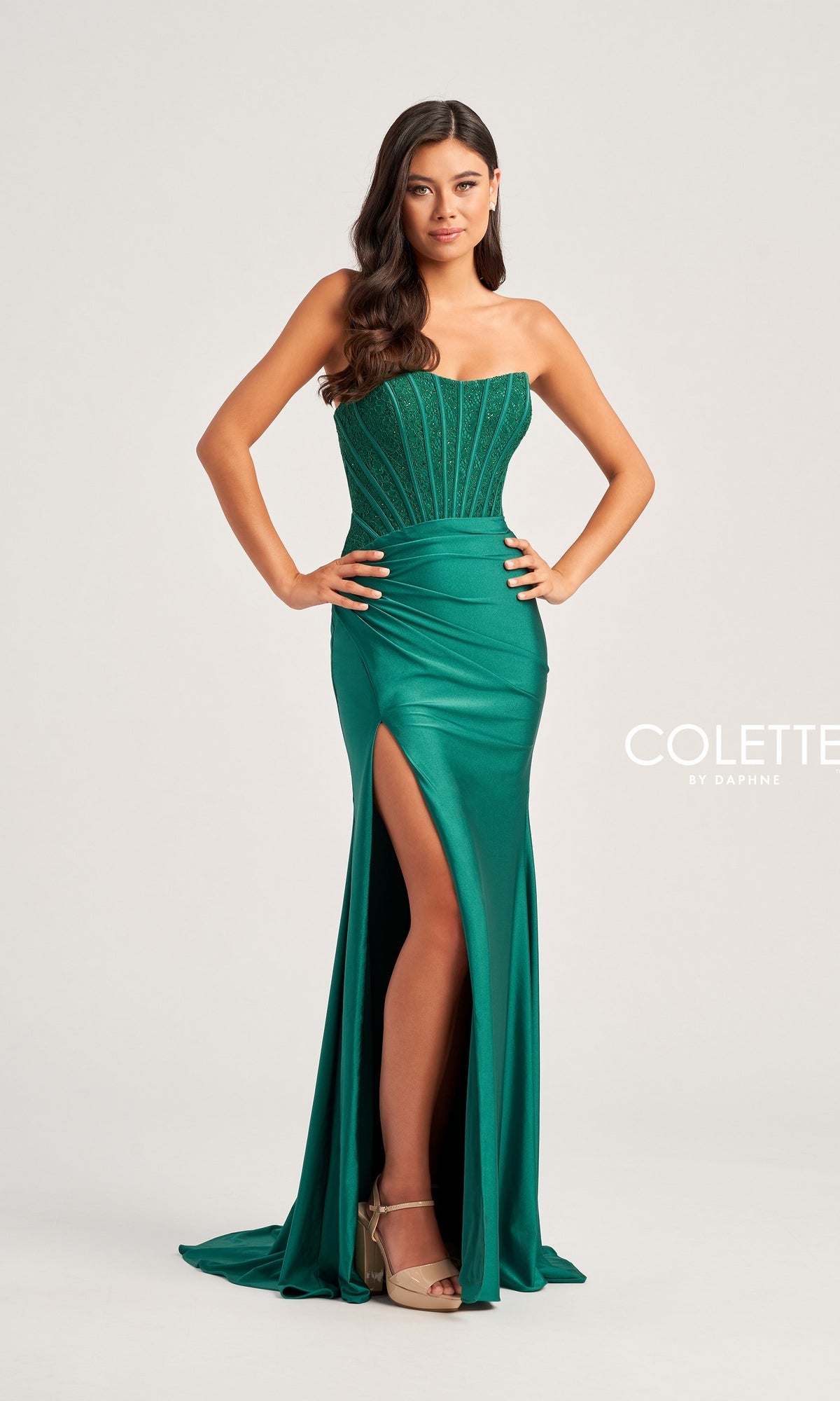 A woman models the Strapless Colette Long Designer Prom Dress CL5158 in emerald green, with a beaded corset bodice and thigh-high slit, standing against a plain background.